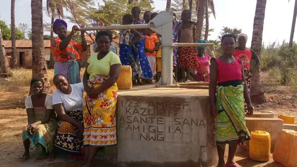 Un pozzo d'acqua in un villaggio del Kenya con la scritta Amref, Asante Sana, Famiglia NWG, con le donne e i bambini del villaggio intorno alla pompa